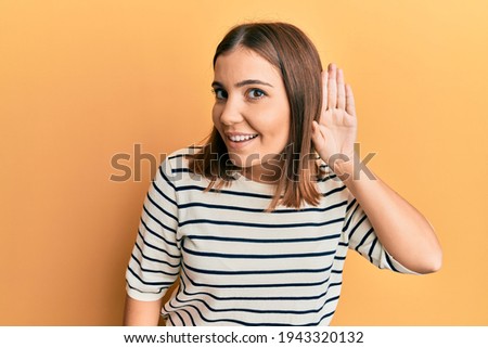 Young beautiful woman wearing casual striped t shirt smiling with hand over ear listening and hearing to rumor or gossip. deafness concept. 