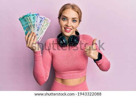 Beautiful blonde caucasian woman wearing gym clothes holding indian rupee smiling happy and positive, thumb up doing excellent and approval sign 