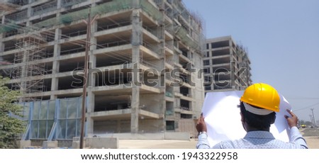 Rear view of an Engineer wearing protective Hard hat and looking through the blueprint of Building Construction ongoing in the Construction site. Businessman . Civil. Royalty-Free Stock Photo #1943322958