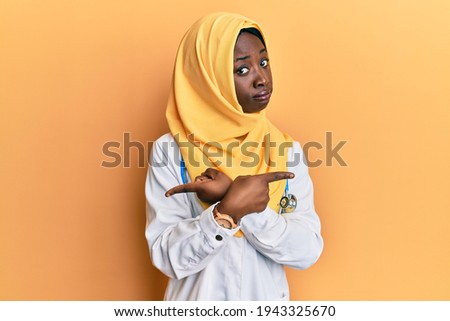 Beautiful african young woman wearing doctor uniform and hijab pointing to both sides with fingers, different direction disagree 