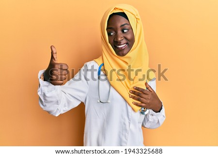 Beautiful african young woman wearing doctor uniform and hijab looking proud, smiling doing thumbs up gesture to the side 