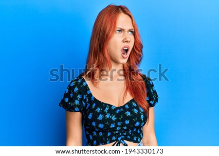 Young beautiful redhead woman wearing casual clothes over blue background angry and mad screaming frustrated and furious, shouting with anger. rage and aggressive concept. 