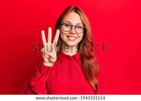 Young beautiful redhead woman wearing casual clothes and glasses over red background showing and pointing up with fingers number three while smiling confident and happy. 