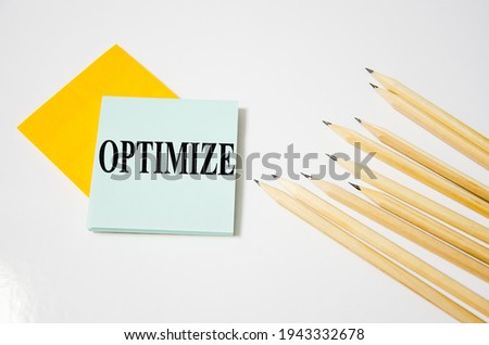 optimize word written on a yellow piece of paper and white background with pencils lying next to it