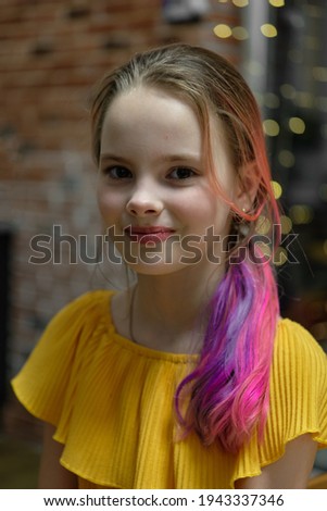A photo of a young girl in a yellow dress, pretty with pink and purple colored hair on Christmas Eve against the background of a glowing garland and a red brick wall.