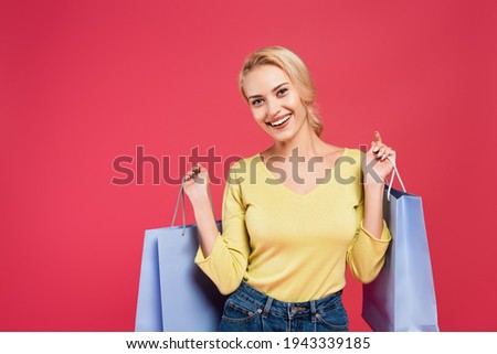 young cheerful woman smiling at camera while holding shopping bags isolated on pink