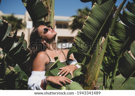 cute young teenage girl or woman posing with banana tree leaves dressed in summer hipster vintage white stylish clothes. Trendy girl posing. Funny and positive woman in sunglasses