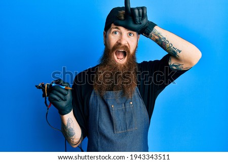 Redhead man with long beard tattoo artist wearing professional uniform and gloves making fun of people with fingers on forehead doing loser gesture mocking and insulting. 