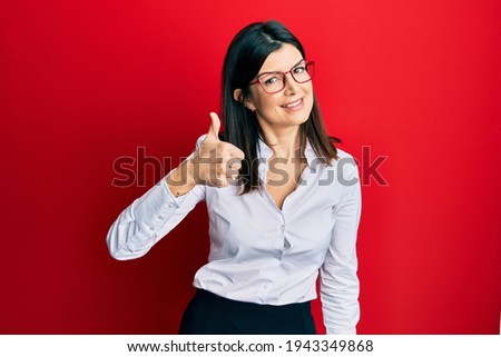 Young hispanic woman wearing business shirt and glasses doing happy thumbs up gesture with hand. approving expression looking at the camera showing success. 