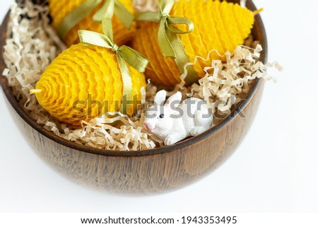 Close up of a gift set of yellow decorative natural beeswax honey candles for interior in the shape of an Easter egg with a ribbon near bunny figurine in a wooden bowl with filler on white background