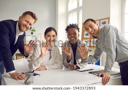 Team of happy young diverse business people sitting and standing around office table, smiling, looking at camera and waving hello, greeting international coworkers in remote work meeting via Internet Royalty-Free Stock Photo #1943372893