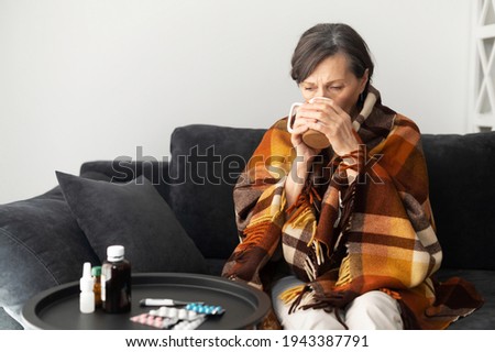 A senior woman feels unwell, sits down on the couch alone at home, covered with blanket feels chill, an elderly woman suffers from viral disease, has fever, treats herself with hot remedy drinks Royalty-Free Stock Photo #1943387791