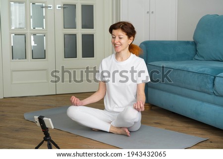 Middle-aged woman with red hair in white clothes, a stream blogger, recording video, looking at the screen of a smartphone, siting on the floor in the living room at home