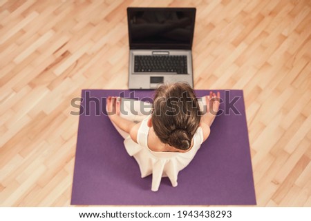 Little girl sitting in the lotus position in front of a laptop. The child trains yoga remotely