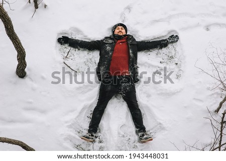 A contented and happy man, a traveler in a black coat and a red sweater, lies, rests on the white snow, like a star, moving his arms like a bird. Photography, concept.