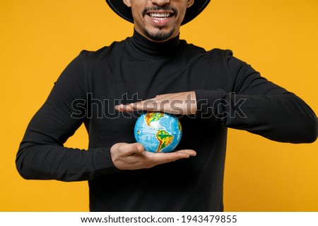 Close up cropped photo shot young geographer fun african man 20s wear stylish black shirt hat eyeglasses holding in palms Earth world globe isolated on yellow orange color background studio portrait.