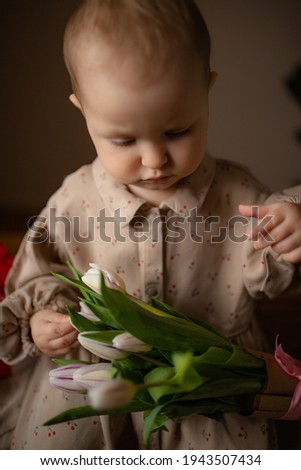 baby girl dressed in beige dress is sitting on a oak wooden table holding a bouquet of purple tulips