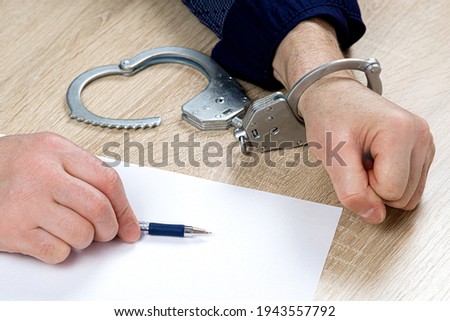 A man writes a confession at the police station. One hand is cuffed, the other is unbuttoned, pen in hand for writing explanations. Arrest, bail, felon, jail. the criminal's fingerprints Royalty-Free Stock Photo #1943557792