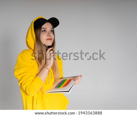Happy woman with a smile in a yellow hoodie and black cap in a notebook draws a rainbow LGBTQ