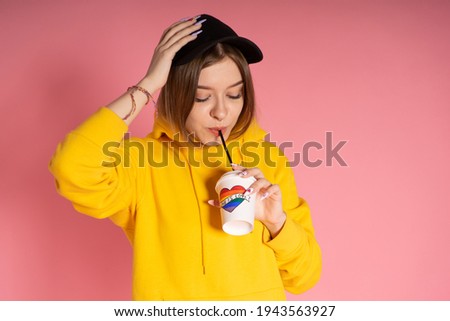 Young LGBTQ woman wearing yellow hoodie jacket drinking coffee from a cup with rainbow heart.