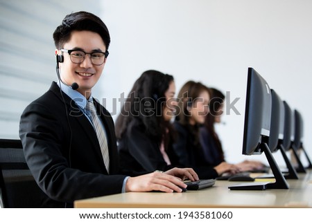 Cheerful Asian man in headset smile with cheerful and happy while working on computer at desk with female coworkers in help desk office. Sevice mind and ready  for marketing support concept.