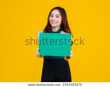 Cute and pretty curly hair Asian female brunette holding green blank board poses to camera with a joyful and positive gesture for advertising use purpose, studio shot isolated on yellow background