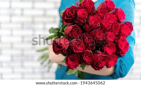 The girl is holding an armful of luxurious red roses on a white brick wall background. Greeting card with a large bouquet of red roses with copy space. Flower delivery banner.