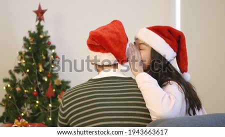 Happy Asian beautiful young family couple husband and wife sitting on sofa back view wearing a santa hat in front fireplace at home Christmas winter hugging giving gift box in living room,