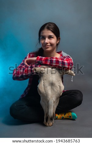 The girl sits on a gray background, resting on the skull of a horned animal