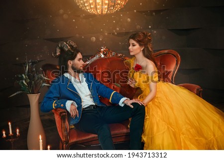 loving couple sits on medieval vintage sofa. Happy beauty woman fantasy princess in yellow dress looks at man prince. guy enchanted beast, horns on head. Blue caftan, tailcoat carnival monster costume Royalty-Free Stock Photo #1943713312