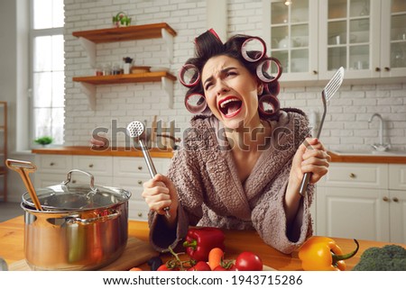 Desperate housewife sick and tired of cooking and housework crying in the kitchen. Unhappy overworked young woman in hair rollers and bathrobe complaining about everyday cooking food and cleaning home Royalty-Free Stock Photo #1943715286
