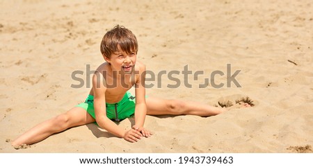 The child in a bathing suit plays with sand at the beach on a sunny day. In the background the beach with people walking by the sea.