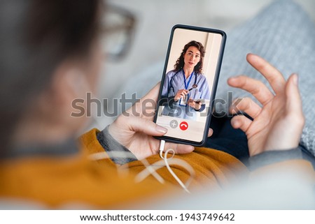 Young woman patient in conversation with specialist over video conferencing using smart phone. Sick woman patient lying on sofa doing video consultation with doctor and explain her symptoms. Royalty-Free Stock Photo #1943749642