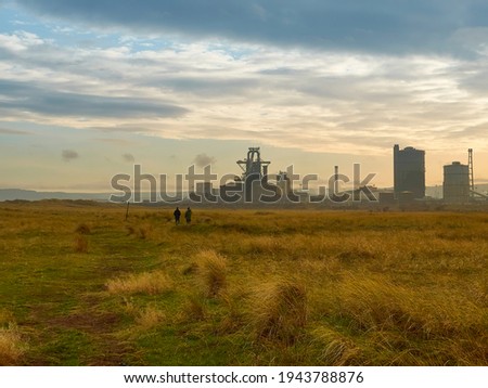 Wide-angle view across the South Gare slag fields, spoil now overgrown with grass. Two people are silhouetted in the distance with the derelict steel mill on the horizon, under a bright winter sky.