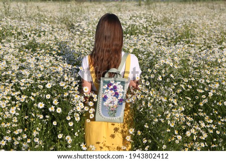 Russian fashionable girl, stylish vintage backpack. Field of wild camomiles. Flower field. Retro style in fashion. Summer day in Russia. July nature, countryside. Russian village. Healthy long hair Royalty-Free Stock Photo #1943802412