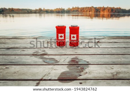 Two cans of beer in red cozy beer can coolers with Canadian flag standing on wooden pier by lake outdoors. Wet footprints on wooden dock. Friends celebrating Canada Day national celebration by water.  Royalty-Free Stock Photo #1943824762