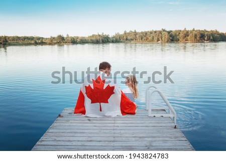 Father and daughter wrapped in large Canadian flag sitting on wooden pier by lake. Canada Day celebration outdoors. Dad and child sitting together on 1 of July celebrating national Canada Day. Royalty-Free Stock Photo #1943824783