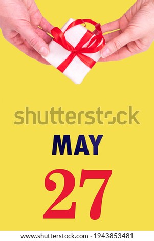 May 27th. Festive Vertical Calendar With Hands Holding White Gift Box With Red Ribbon And Calendar Date 27 May On Illuminating Yellow Background. Spring month, day of the year concept.