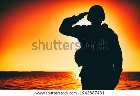 Silhouette of soldier in combat helmet and ammunition saluting on background of sunset sky. Army special forces fighter, Marines rifleman showing respect, greeting officer with salute gesture Royalty-Free Stock Photo #1943867431