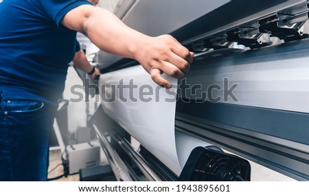 man holding printable material on alarge format printing plotter.graphic design and advertising concept. Royalty-Free Stock Photo #1943895601