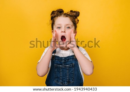 Portrait of little preschool girl call somebody hold hands near face, isolated on yellow studio background, small pretty kid open mouth look at camera shout speak loud make announcement concept Royalty-Free Stock Photo #1943940433
