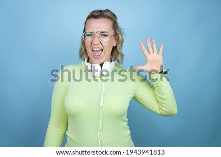 Young caucasian woman wearing headphones on neck over blue background showing and pointing up with fingers number five while smiling confident and happy