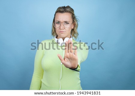 Young caucasian woman wearing headphones on neck over blue background serious and doing stop sing with palm of the hand.
