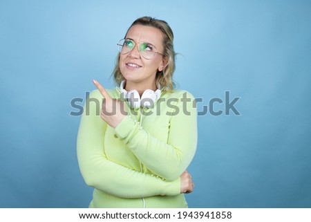 Young caucasian woman wearing headphones on neck over blue background confused and pointing with hand and finger to the side