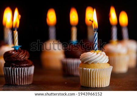Close up side view image of an assortment of chocolate and vanilla cupcakes with white and brown frostings on. Each have a candle lit against dark background. Celebration, party, birthday concepts.
