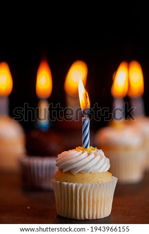 Close up side view image of an assortment of chocolate and vanilla cupcakes with white and brown frostings on. Each have a candle lit against dark background. Celebration, party, birthday concepts.