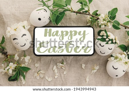 Happy Easter text on phone screen with easter eggs with cute faces in floral wreaths on rustic fabric with blossoms, handwritten sign. Beautiful stylish greeting card. Seasons greetings