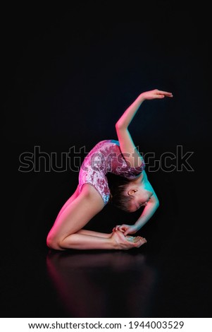 Young flexible girl isolated on a black studio background in neon light. A young female model is engaged in rhythmic gymnastics. Exercises for flexibility, balance Grace in movement, sport, and action
