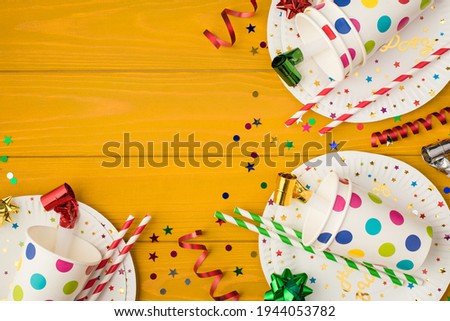 Overhead photo of cups drink with party tubules whistle multicolored confetti bow and plates isolated on the yellow wooden background with empty space