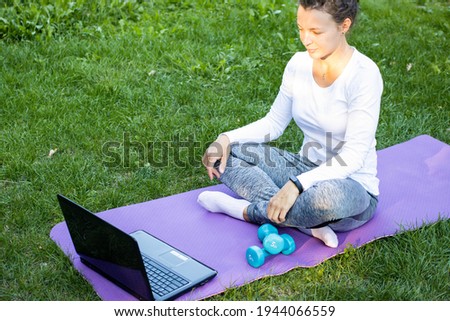 Young fit woman watching online fitness class at laptop screen or communicate with personal trainer in distant, sitting on sport mat in green park outdoor, using wireless 5g internet connection.